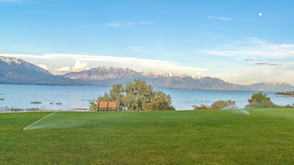 Wall Mural - Pano Sprinklers on vast grassy terrain with blue lake snowy mountain and sky view