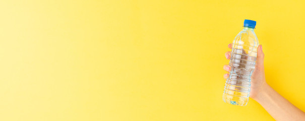 Female hand holding bottle of water on yellow background with copyspace. Banner