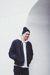 Wall Mural - stylish laughing guy in a black hat and winter jacket near a white wall