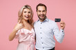 © ViDi Studio - Funny young couple two guy girl in party outfit celebrating posing isolated on pastel pink background. Valentine's Day, Women's Day birthday holiday concept. Hold credit bank card, showing thumb up.