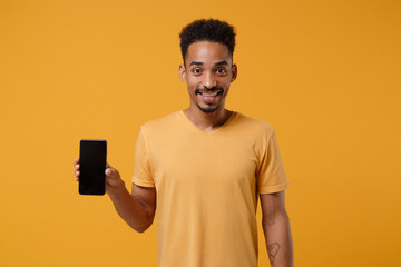 Wall Mural - Smiling young african american guy in casual t-shirt posing isolated on yellow orange background studio portrait. People lifestyle concept. Mock up copy space. Holding mobile phone with blank screen.
