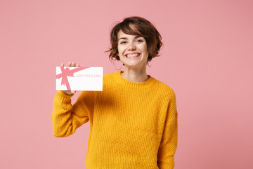 Cheerful young brunette woman girl in yellow sweater posing isolated on pastel pink background studio portrait. People sincere emotions lifestyle concept. Mock up copy space. Holding gift certificate.