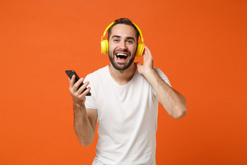 Laughing young man in casual white t-shirt posing isolated on orange wall background studio portrait. People lifestyle concept. Mock up copy space. Listening music with headphones, hold mobile phone.