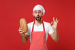Shocked young bearded male chef cook or baker man in striped apron white t-shirt toque chefs hat isolated on red background. Cooking food concept. Mock up copy space. Holding bread showing OK gesture.