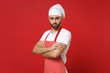 Handsome young bearded male chef cook baker man in striped apron white t-shirt toque chefs hat posing isolated on red wall background. Cooking food concept. Mock up copy space. Holding hands crossed.