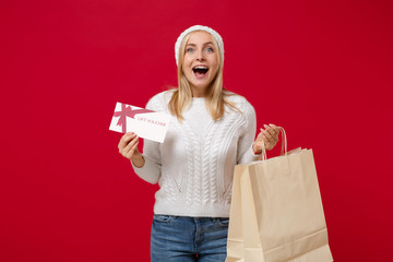 Wall Mural - Excited young woman in sweater, hat isolated on red background. Healthy fashion lifestyle, cold season concept. Mock up copy space. Hold package bag with purchases after shopping, gift certificate.