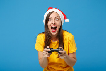 Poster - Excited young woman Santa girl in yellow t-shirt Christmas hat posing isolated on bright blue background. Happy New Year 2020 celebration holiday concept. Mock up copy space. Hold joystick play game.