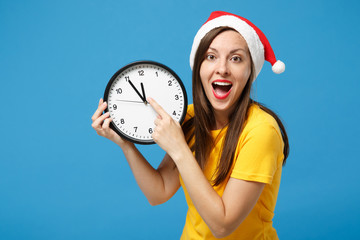 Wall Mural - Funny young brunette woman Santa girl in yellow t-shirt Christmas hat posing isolated on blue background. New Year 2020 celebration holiday concept. Mock up copy space. Pointing index finger on clock.