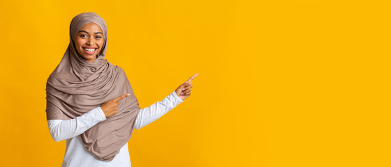 Wall Mural - Smiling black muslim woman pointing at copy space on yellow background