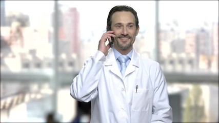 Wall Mural - Happy caucasian pharmacist talking on phone. Cheerful middle-aged man in white robe using smartphone on blurred background.