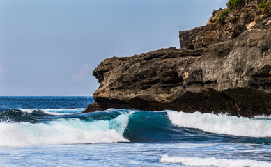 Wall Mural - Waves at Secret Beach, Nusa Ceningan, Bali, Indonsia. Rocky cliff in background. Blue sky in distance. 