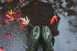 Feet in olive green rubber boots standing in a puddle with fallen leaves.