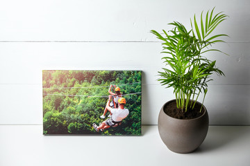 Canvas print on white table, front view. Photo with gallery wrap method of canvas stretching on stretcher bar. House plant and a color photography with image of people on zip line. Interior decor