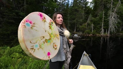 Wall Mural - A spiritual caucasian woman seeks mindfulness and enlightenment in nature, playing a native shamanic handcrafted drum and beater by a calm lake