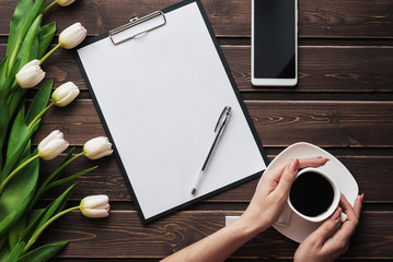 Wall Mural - White tulips on a wooden table with an empty paper tablet and smartphone and a Cup of coffee in women's hands