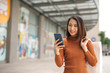© makistock - Portrait of elegant young Asian woman holding shopping bags and using smartphone on the go while leaving mall