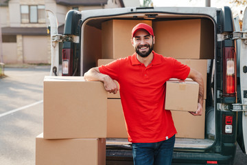 Wall Mural - Image of joyful young delivery man standing with parcel boxes near car