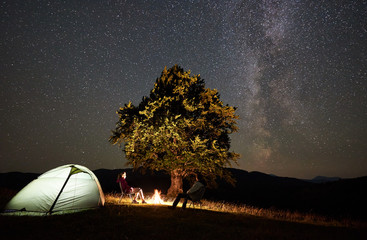 young family hikers relaxing at summer night camping in mountains. man and woman sitting on chairs b