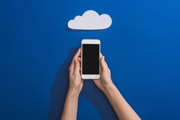 Wall Mural - cropped view of woman holding smartphone near empty white paper cloud on blue