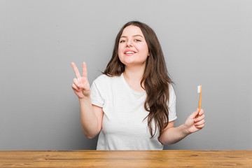 Wall Mural - Young plus size curvy woman holding a toothbrush showing number two with fingers.