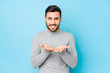 © Asier - Young caucasian man against a blue background isolated holding something with palms, offering to camera.