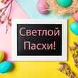 © fascinadora - Orthodox Easter concept. Colorful eggs and pussy willow branches on bright colorful background and chalkboard with russian letters Happy Easter. Top down view or flat lay. Square crop for social media