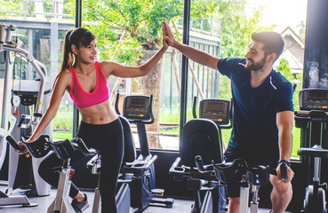 Young sporty woman and man giving each other a high five after cycling training in gym. Fit couple high five after workout in health club, Fitness class doing sport biking in the gym for health. .