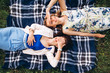 Two pretty girls lie on a blue plaid outdoors in the park, summer picnic. Girlfriends' girls have fun, laugh, smile, and frolic. Closeup portrait of two young women in dresses and straw hats.