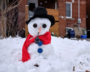 Snowman dressed with red scarf black felt hat blue buttons smoking a pipe in front of blurred residence