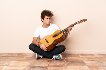 Wall Mural - Young caucasian man with a guitar sitting on the floor with sad expression