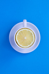 Wall Mural - White tea cup with fresh halved lemon in middle on the blue background. Top view. Copy space. Creative food collage. Location vertical.