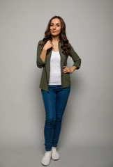 Wall Mural - Perfect hair. Full-length photo of a dark-haired woman in a khaki shirt and blue jeans, who is posing in front with her left hand on her hip, looking in the camera and smiling.