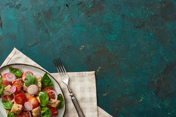 Wall Mural - top view of delicious Italian vegetable salad panzanella served on plate on textured green surface with napkin and fork