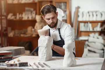 Wall Mural - Male sculptor in white shirt and black apron makes a limestone copy of woman torso at the artistic studio.