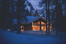 A Cozy Wooden Cabin Cottage Chalet House Covered In Snow Near Ski Resort In Winter With The Lights Turn On