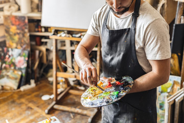 Male painter at art studio indoors mixing colors on palette with brush pensive working process close-up