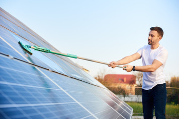 Young man is washing a solar panel with a mop, pv plant in rural area, cleaning increases to high performance, side view, close-up