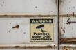 a faded Premises Under 24hr Surveillance warning sign on the door of a disused industrial building
