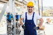 Full length of handsome caucasian unshaven smiling worker in overall and with helmet on head posing next to dashboard in refinery.