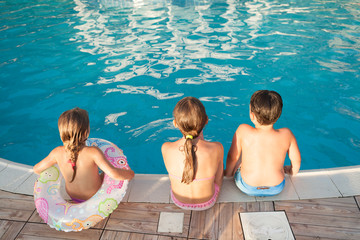 Two children and a girl with a rubber ring are sitting on the edge of the pool.