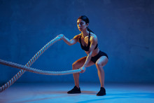 Caucasian Young Female Athlete Practicing On Blue Studio Background In Neon Light. Sportive Model Training Her Upper Body With Ropes. Body Building, Healthy Lifestyle, Beauty And Action Concept.