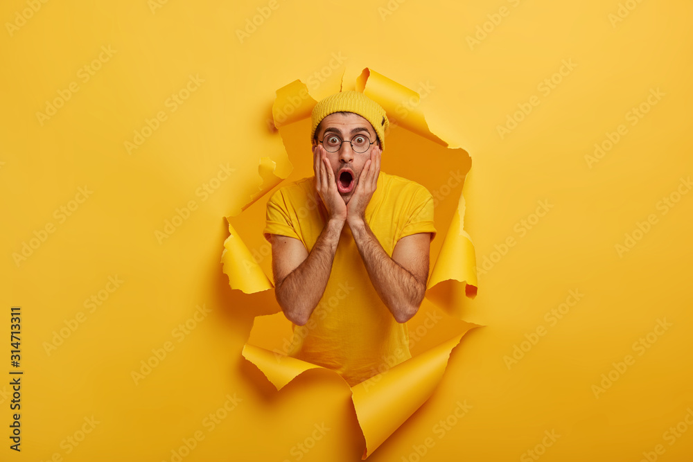Speecheless impressed guy keeps hands on cheeks, stares at camera with opened mouth, gossips about something, wears yellow casual clothes, stands in ripped hole background. Emotions concept. - obrazy, fototapety, plakaty 