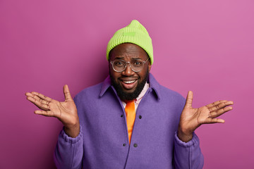 Wall Mural - Portrait of dark skinned guy has no clue what happening, spreads palms, wears optical glasses, hat and purple jacket, has puzzled cheerful expression poses indoor. People, reaction, facial expressions