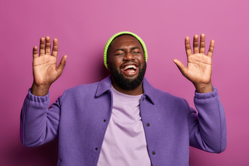 Wall Mural - Joyful bearded Afro American man raises palms, cannot stop laughing, hears funny anecdote, feels overemotive, watches hilarious comedy or joke, dressed in purple coat and green hat, models indoor