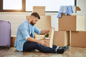 Caucasian man wear blue shirt sit on floor, unbox in new house. Background moving boxes, suitcase, window. The sun shine