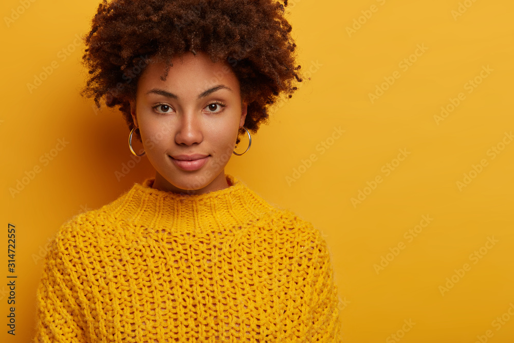 Portrait of Afro American millennial woman with calm serious face expression, looks directly at camera, wears knitted jumper, has natural beauty, models against yellow studio wall with empty space - obrazy, fototapety, plakaty 
