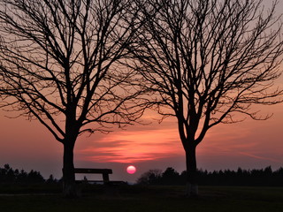 A moody sunset in Franconia with the sun shining shortly above the horizon between two bare trees and besides a bench. Seen in March near the town of Neunhof in Franconia / Bavaria, Germany