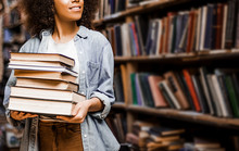 Photo For Background, Place For Insertion, African American Woman Student Holding A Heavy Armful Of Books In Her Hands Against The Background Of Bookshelves In A Library. Learning Concept, Lifestyle