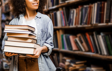 Photo for background, place for insertion, African American woman student holding a heavy armful of books in her hands against the background of bookshelves in a library. Learning concept, lifestyle