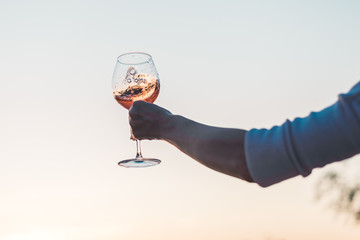 Wall Mural - Glass of rose wine with splashes in woman's hand against the sunset sky.